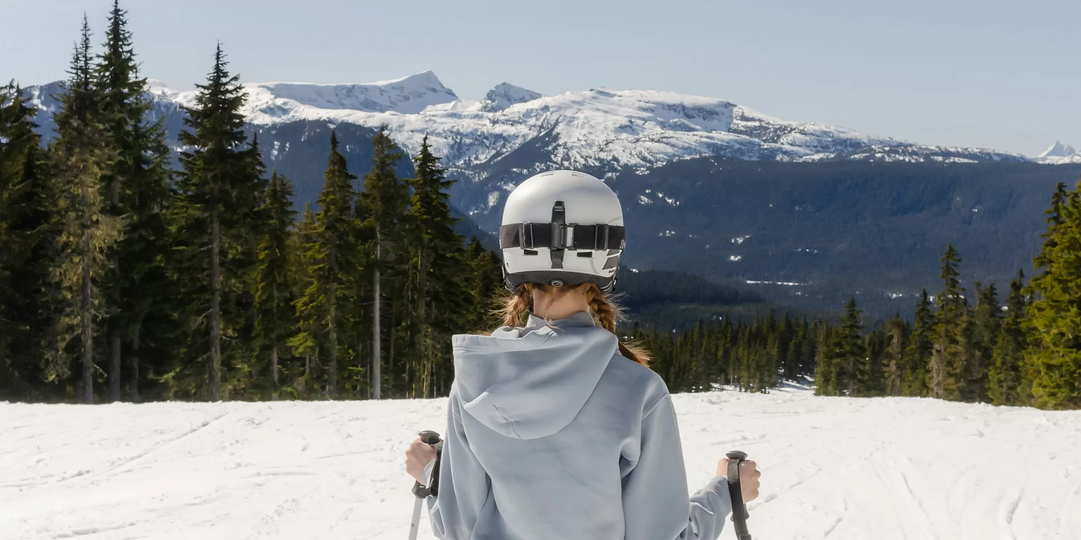 Person wearing a helmet and holding ski poles, standing on a snow-covered slope. They face a scenic view of forested hills and snow-capped mountains under a clear sky.