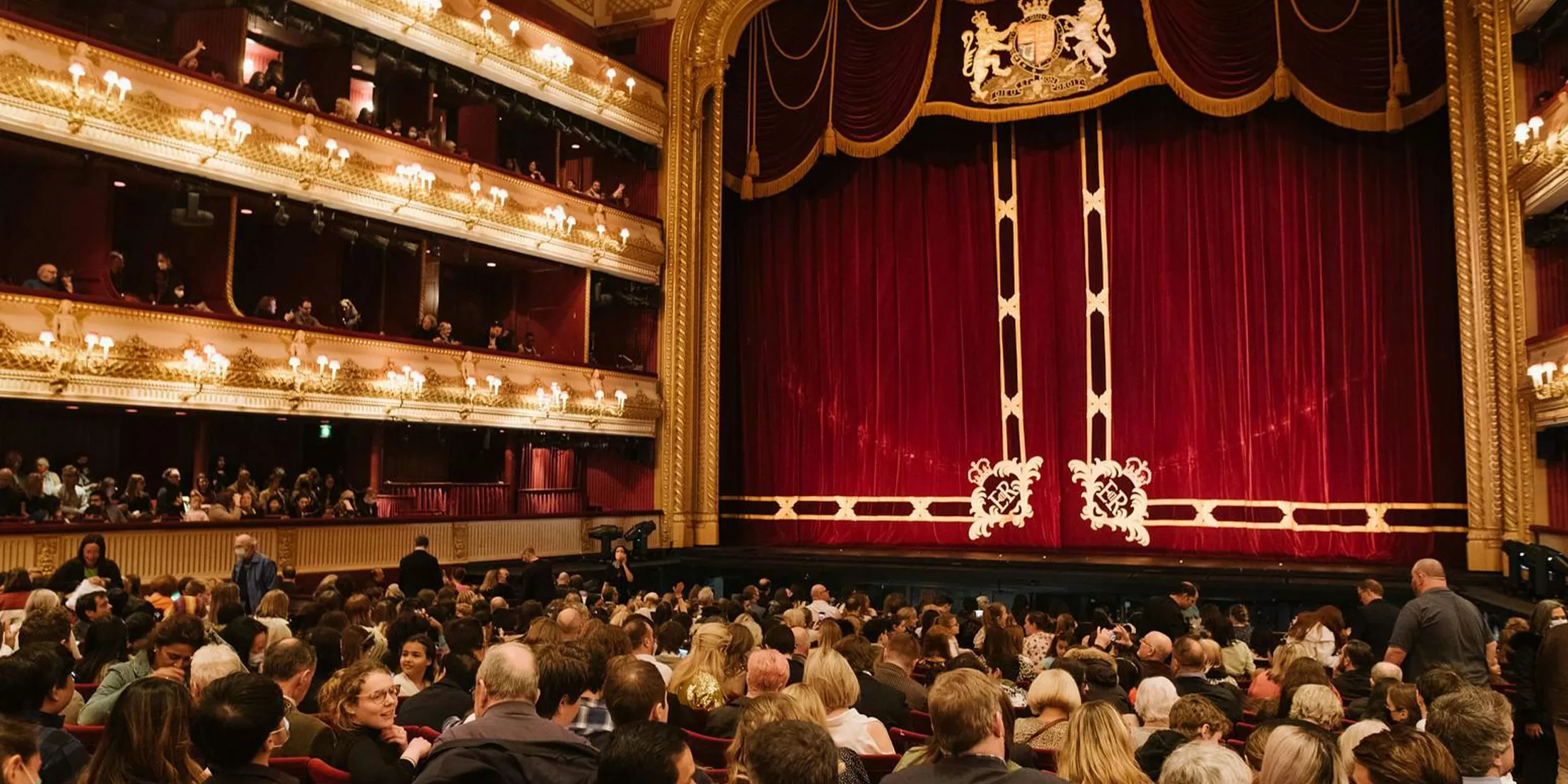 A large audience fills the seats of an ornate theater with golden balconies, elegant lighting, and a grand red curtain on stage, waiting for a performance to begin.
