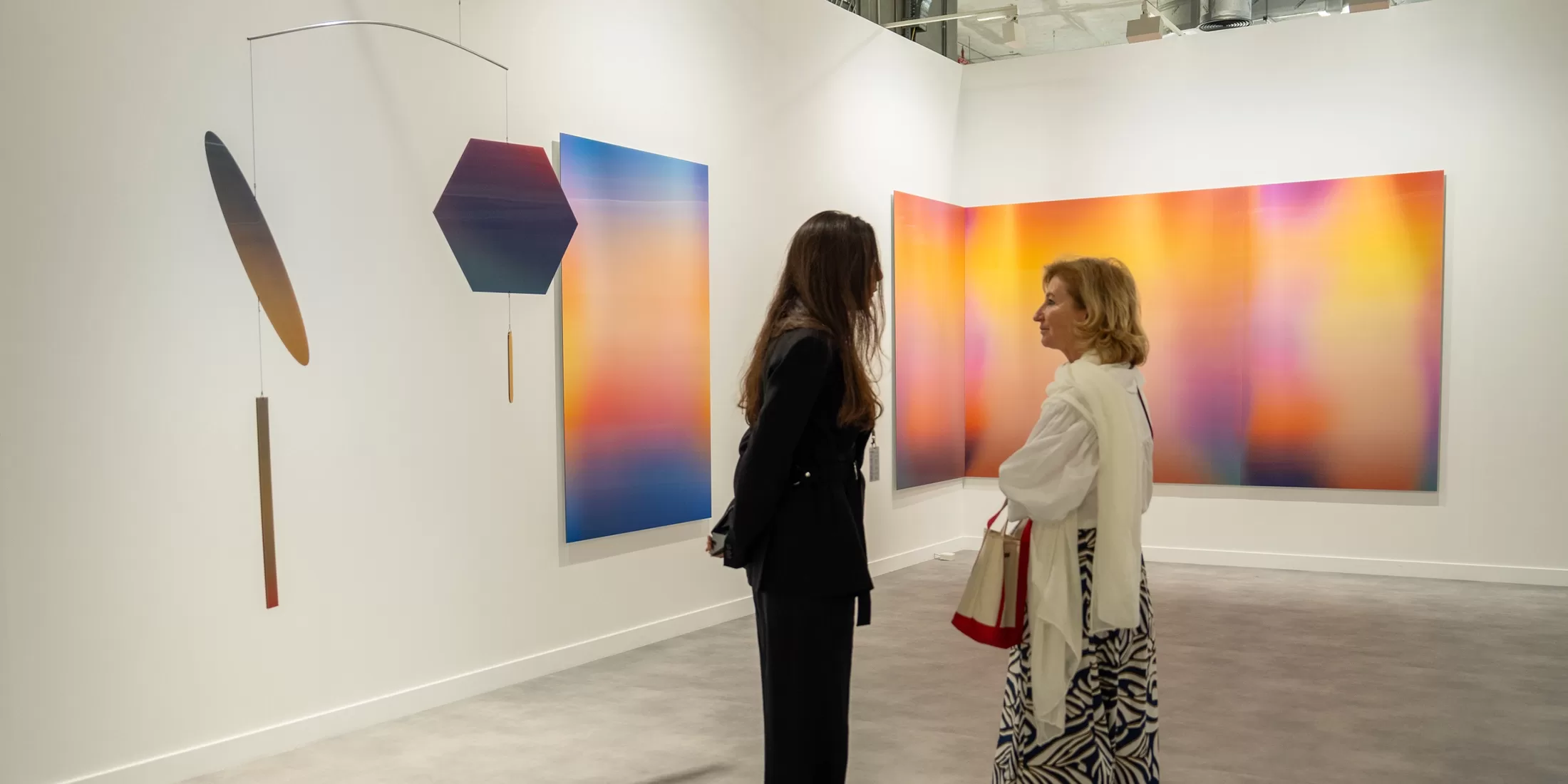 Two women stand and converse in a modern art gallery, surrounded by colorful gradient paintings and a geometric mobile hanging from the ceiling. The space is bright with white walls and soft lighting.