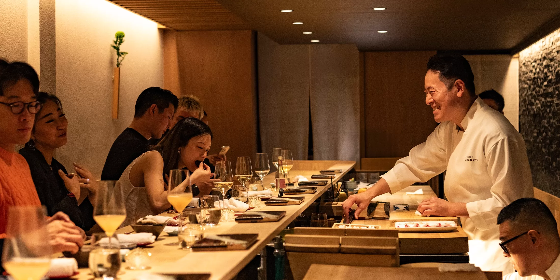 A sushi chef smiles and serves sushi to customers seated at a wooden counter in a warmly lit, modern restaurant. Diners enjoy their food and drinks, creating a lively and inviting atmosphere.