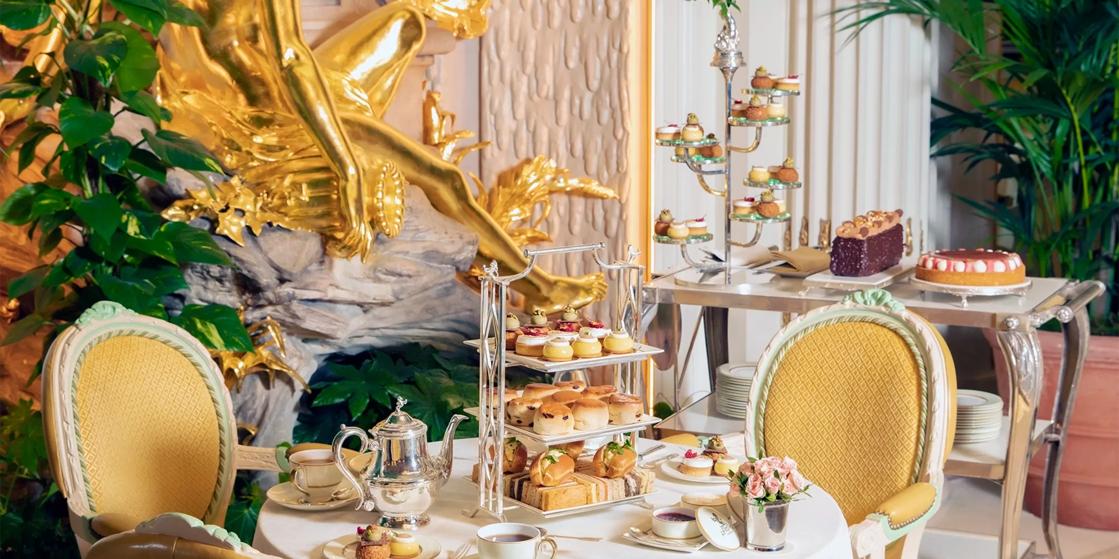 A grand looking room with green leaves and a golden cherub coming from the walls. There is 2 laid tables next to each other. Both tables are filled with multiple variations of scones, cakes and jam. There is a silver tea pot and cups of tea on one table.