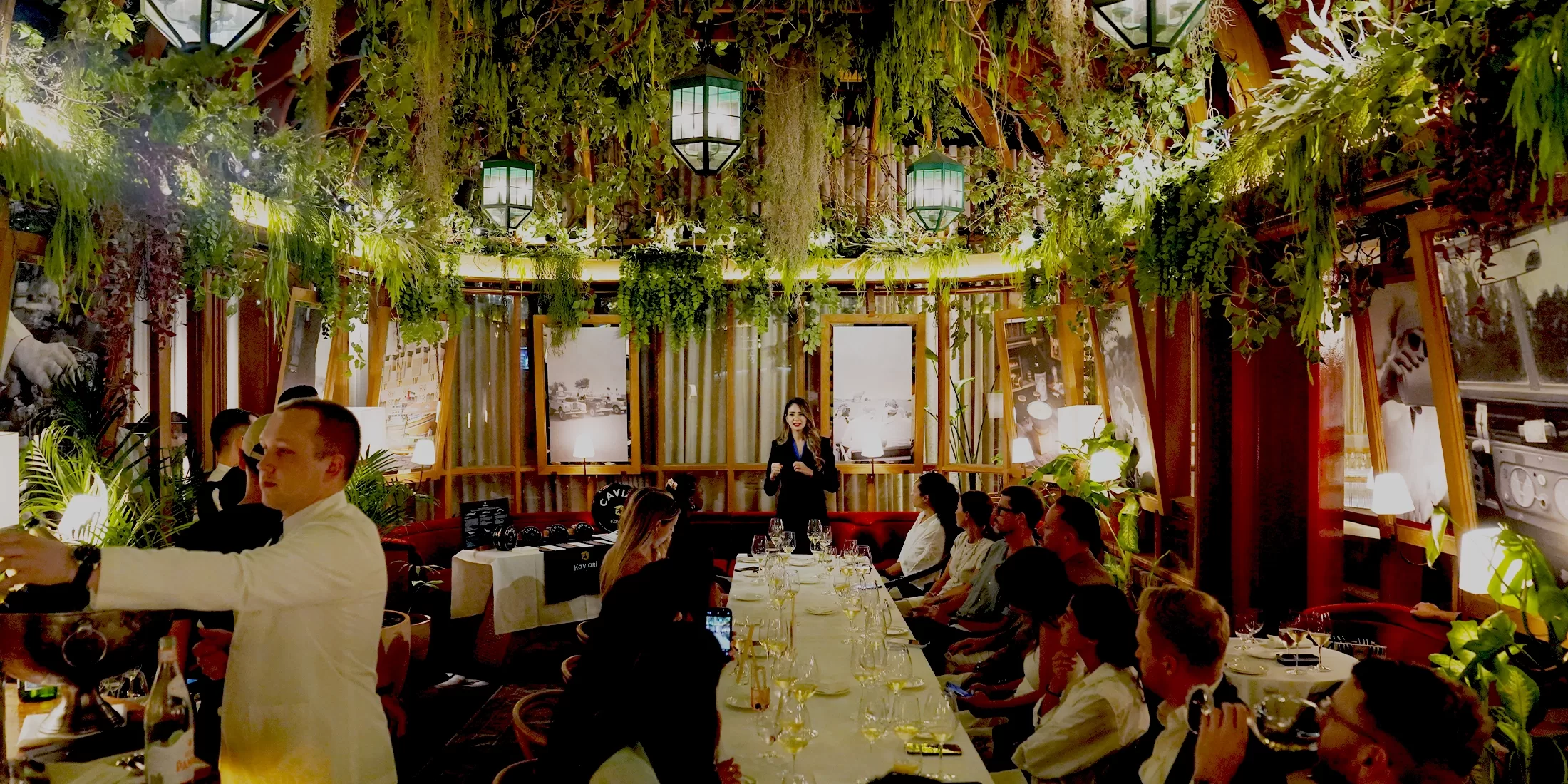 A bustling restaurant scene featuring patrons seated at long tables, enjoying their meals and engaging in conversation whilst a female stands up to present.