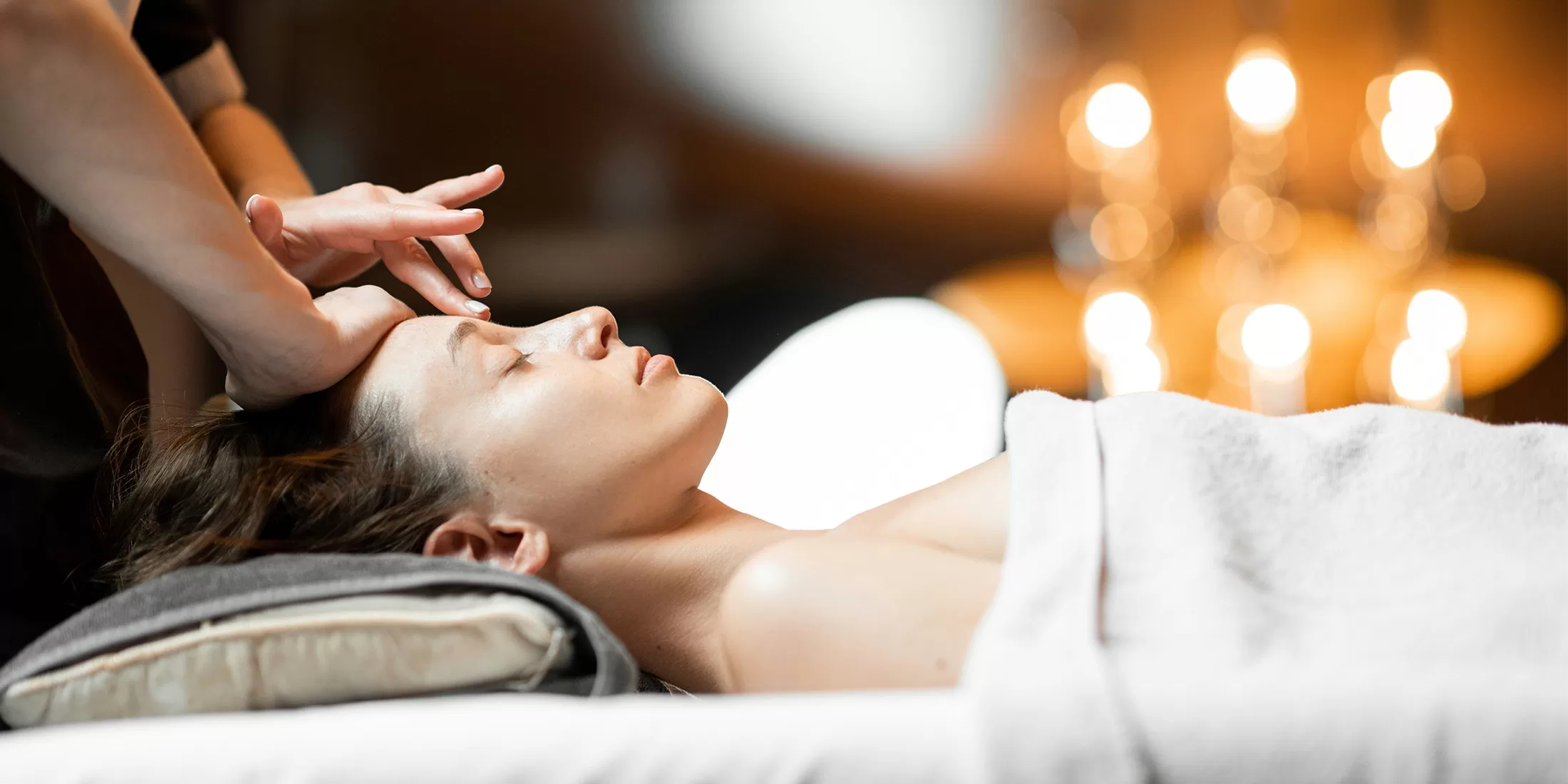 A person lying on a massage table with eyes closed, covered by a white towel. A therapist gently massages their forehead. The setting is softly lit, with blurred warm lights in the background, creating a relaxing ambiance.