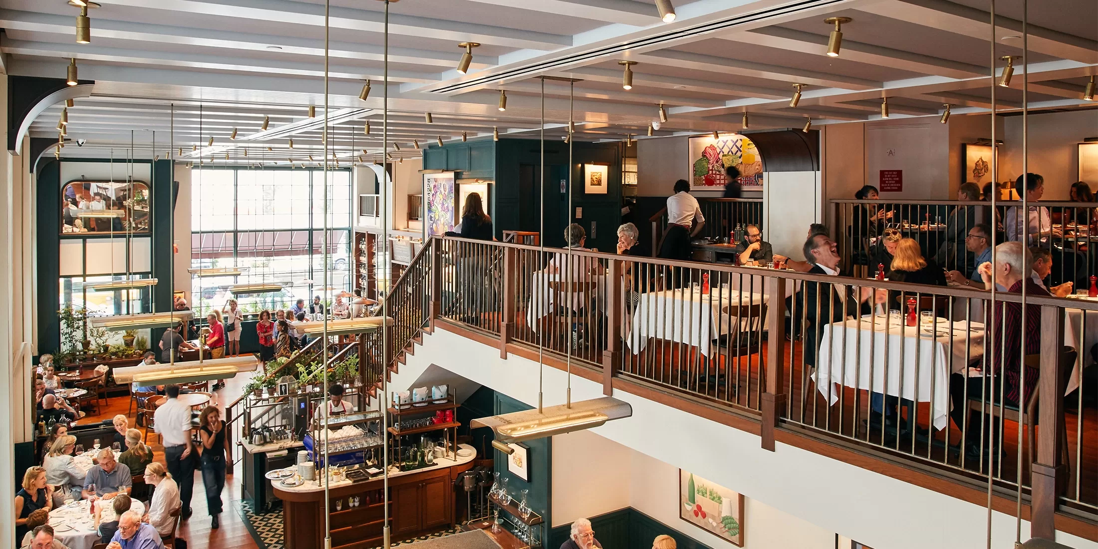 A bustling two-story restaurant with diners seated at tables enjoying meals. The upper level features people sitting at tables with white tablecloths, while downstairs is a vibrant bar area. Large windows let in natural light, enhancing the lively atmosphere.