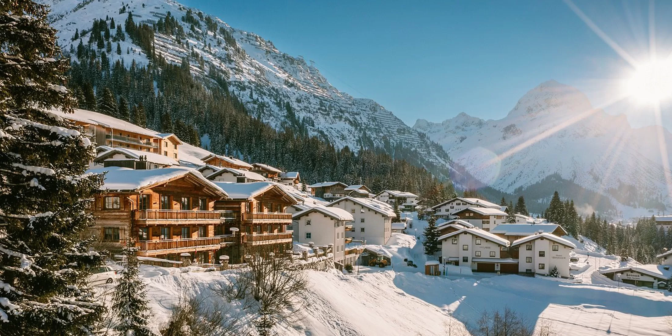 A picturesque mountain village with wooden chalets and snow-covered rooftops, nestled among tall evergreen trees. Majestic snow-capped peaks rise in the background under a clear blue sky, with the sun shining brightly.