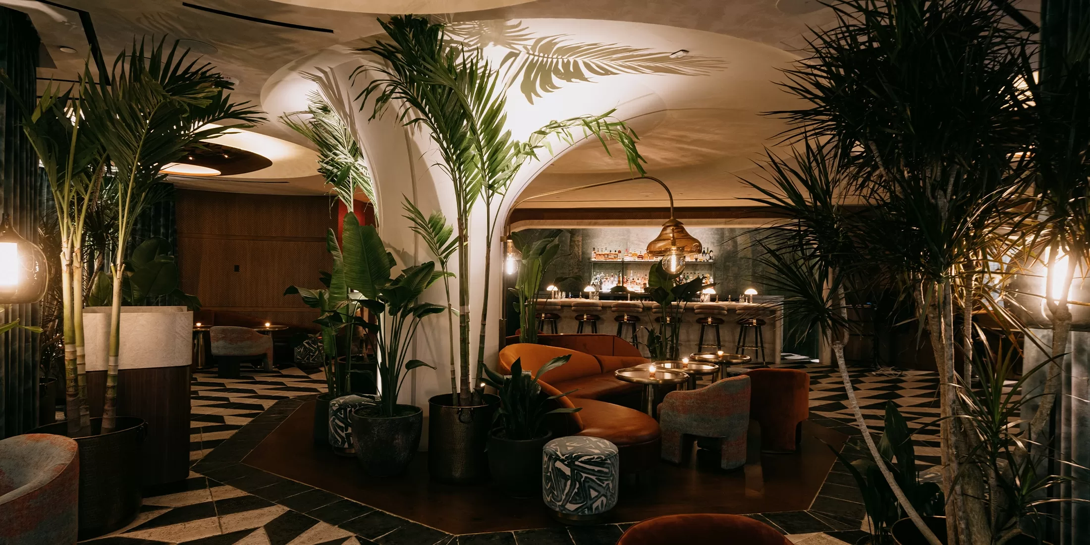 A stylish bar area with tropical plants and warm ambient lighting. Curved white pillars and a mix of modern and vintage furniture create a cozy, elegant atmosphere. The patterned floor complements the lush, leafy decor.