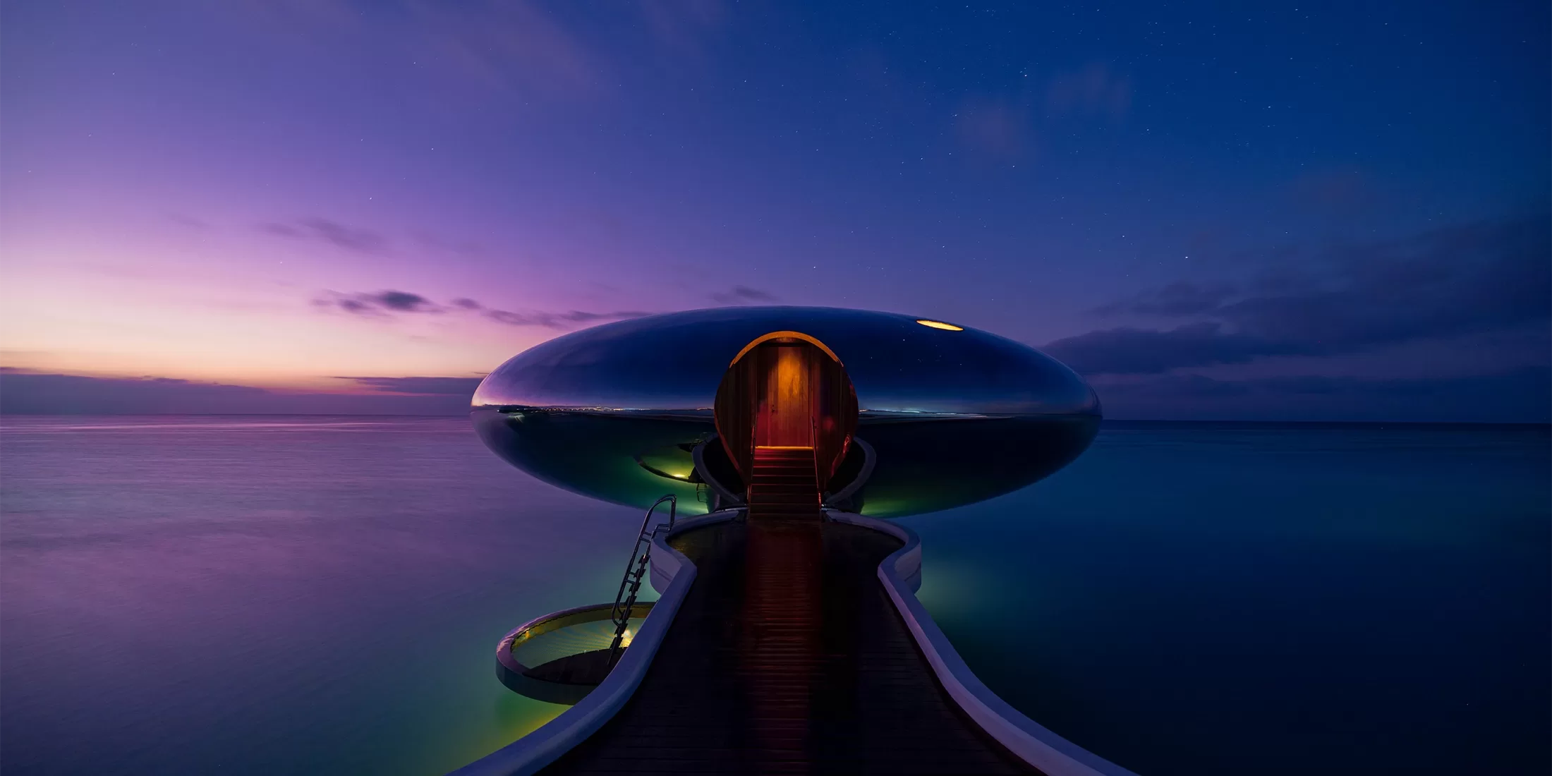 A futuristic, oval-shaped structure glows with ambient lighting over calm ocean waters at dusk. A wooden walkway leads to the entrance, with skies painted in serene shades of purple and blue as stars begin to appear.