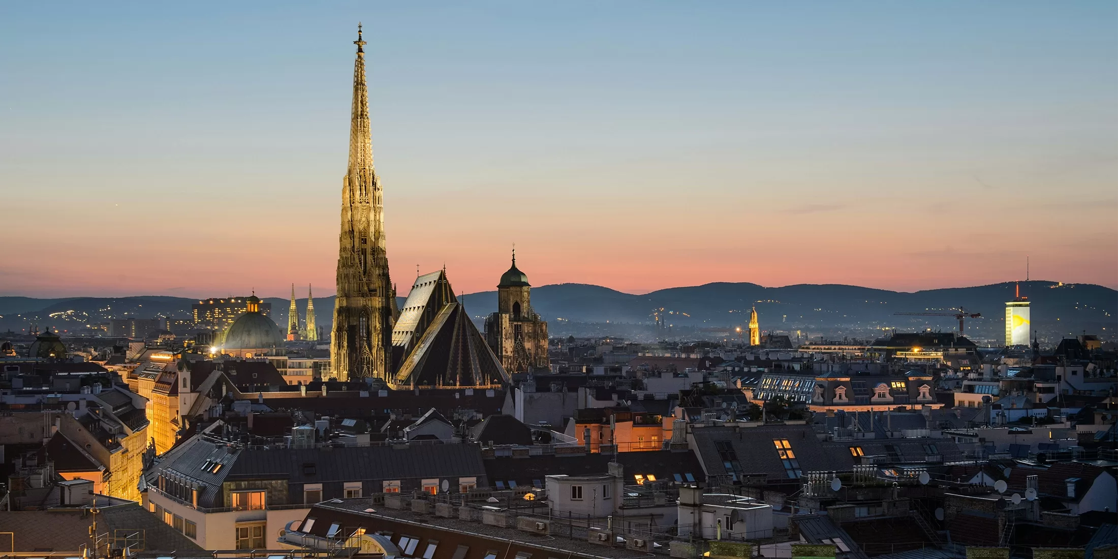 A panoramic view of Vienna at dusk features St. Stephens Cathedral prominently against a pastel sky. City lights begin to twinkle among rooftops, with distant hills silhouetted against the fading light.