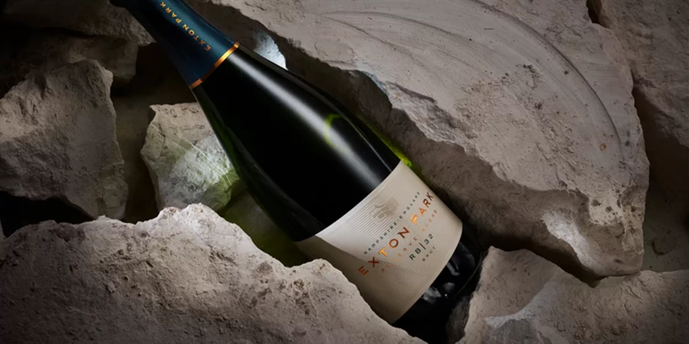 A bottle of Exton Park sparkling wine rests in a rugged rock crevice. The label is visible, displaying the brand name and vintage year. The setting contrasts the dark bottle with the rough, textured stone.