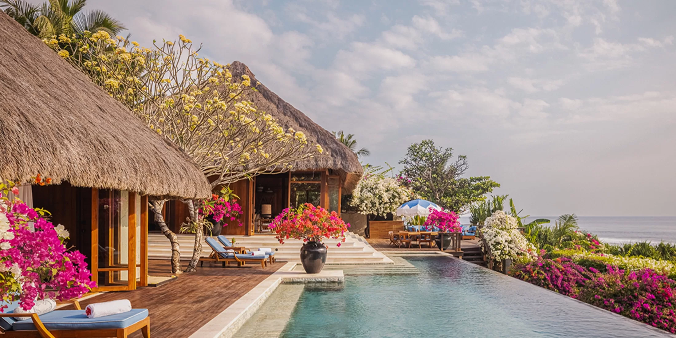 Tropical beachfront villa with thatched roofs, a long pool, and vibrant flowers. Sun loungers are placed around the pool, overlooking the ocean under a partly cloudy sky. Lush greenery surrounds the serene setting.