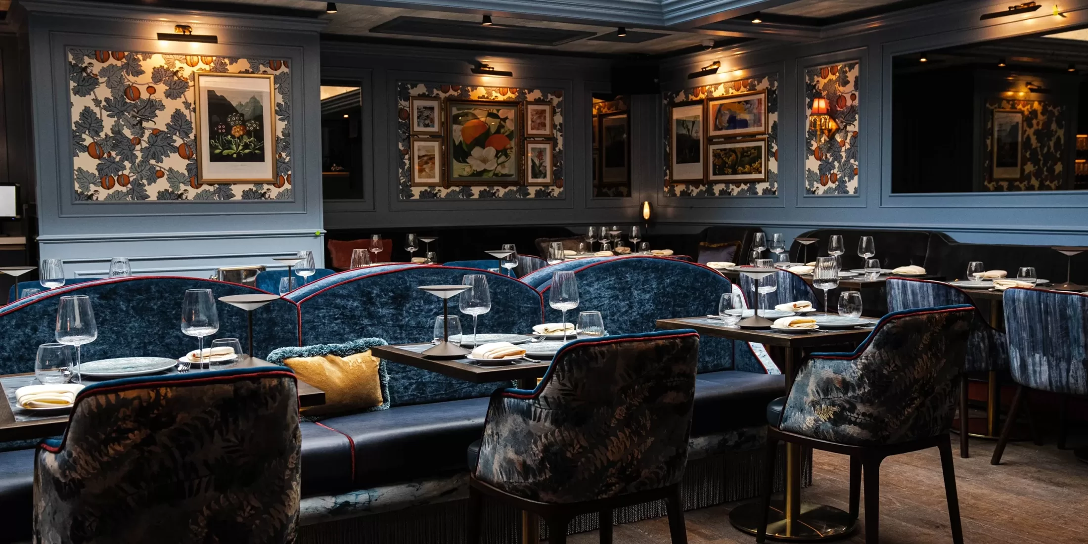 A stylish restaurant interior with dark blue velvet seating and decorative wallpaper. Tables are set with glassware and plates, and framed artwork adorns the walls. Warm lighting provides an elegant ambiance.