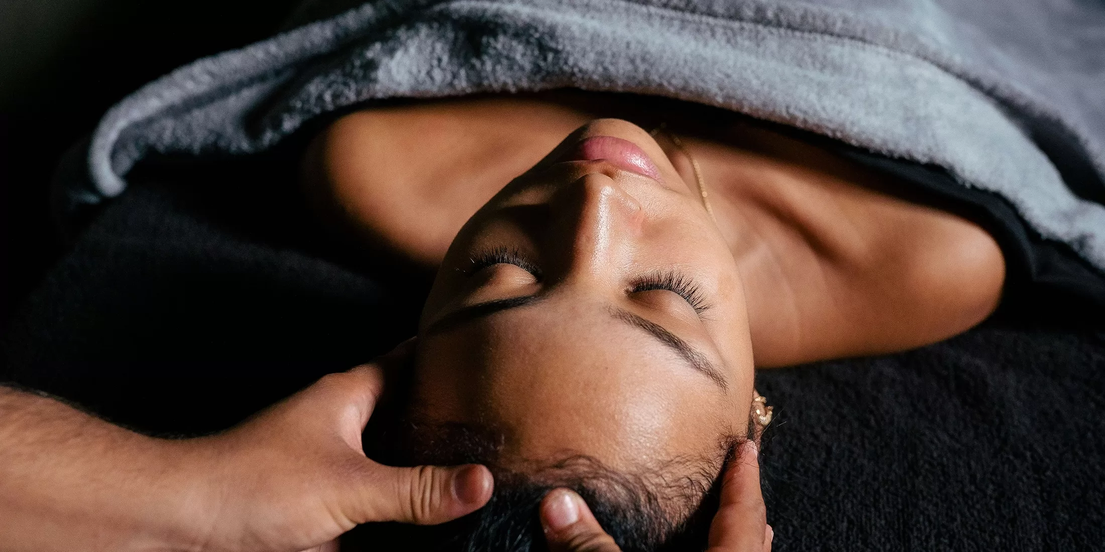 A person lying down with eyes closed, draped in a gray towel, receiving a head massage. Hands gently cradle their head, creating a relaxing atmosphere.