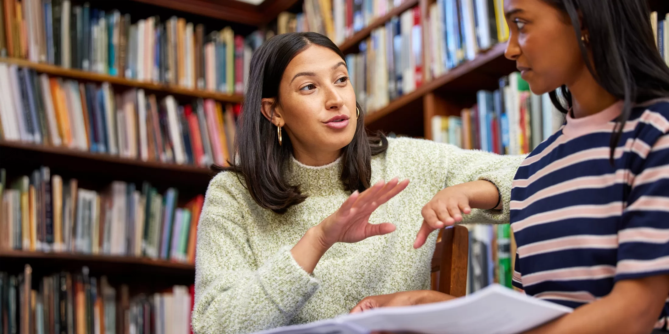 Two women are sitting and talking in a library filled with bookshelves. One woman is gesturing with her hand while holding papers, and both seem engaged in conversation.