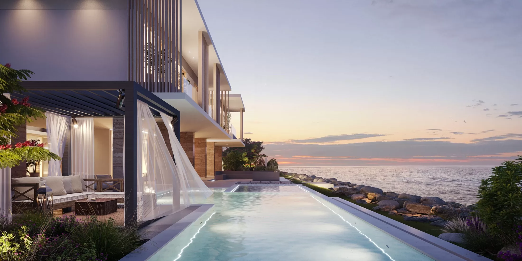 Modern beachfront house at sunset with large glass windows, outdoor seating area, and an infinity pool overlooking the ocean. The sky is painted with soft pastel hues, and the scene exudes tranquility and luxury.