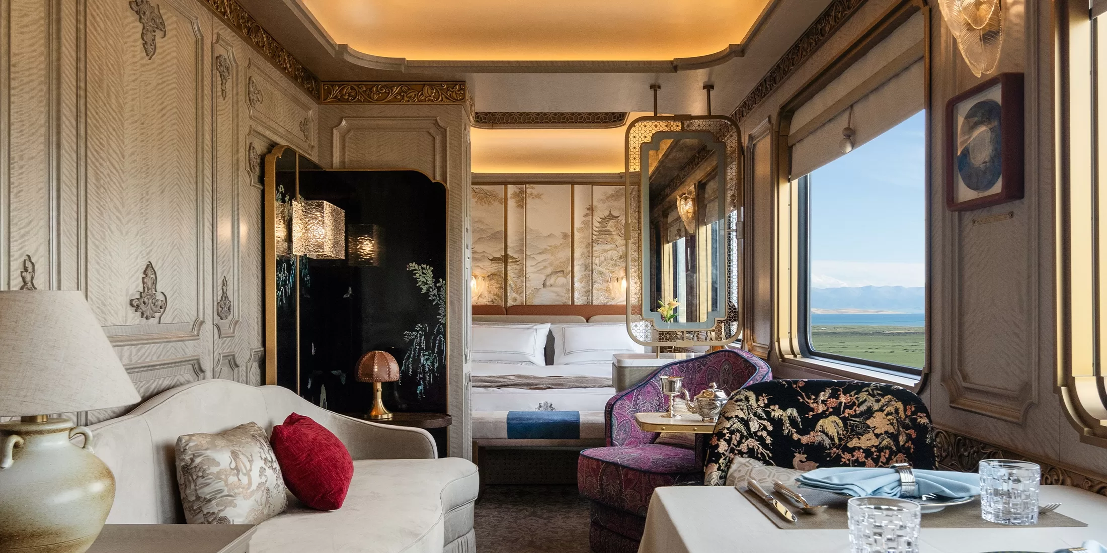 Luxurious train compartment with elegant decor, including a plush sofa, ornate lamp, and dining table set. Large windows reveal scenic mountain and sky views. A cozy bed with pillows is visible in the background.