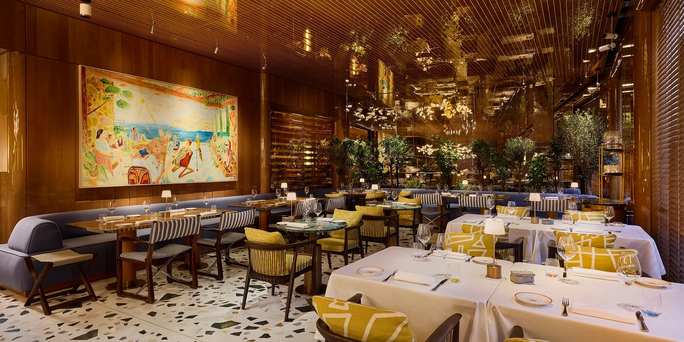 A luxurious restaurant with elegant decor, featuring a large mural of a beach scene on the wall. The room has a warm, golden hue with wooden accents, plush seating, and tables set with white cloths and fine dining ware. Lush greenery adds to the ambiance.