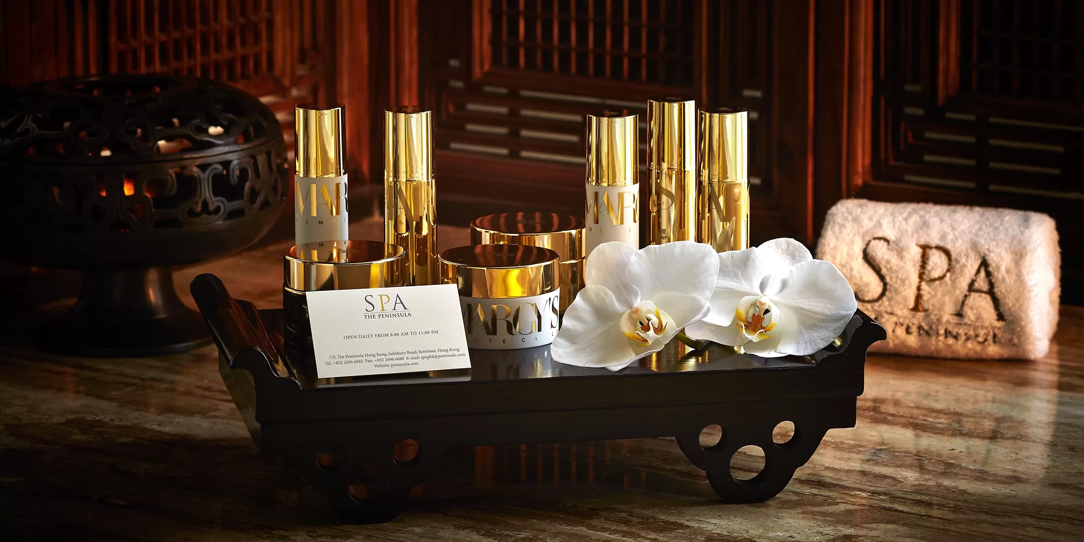 A wooden tray holds luxurious spa products with gold accents, accompanied by white orchids. A candle holder glows in the background, and a rolled towel with SPA embroidered is beside the tray, creating an elegant spa ambiance.