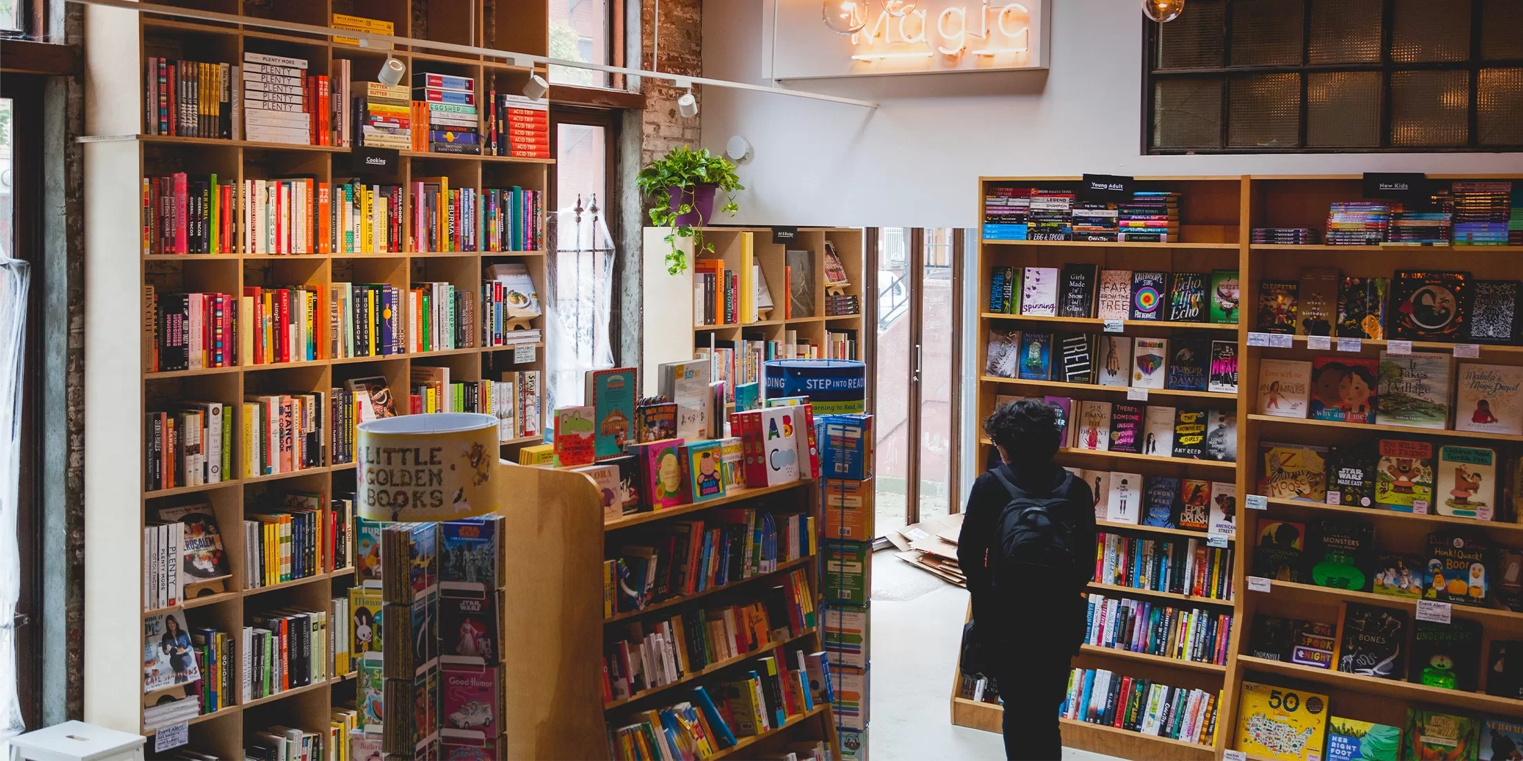 A person stands in a cozy bookstore, surrounded by tall wooden shelves filled with colorful books. A neon sign reading magic hangs on the wall. Soft light filters through large windows, creating a warm and inviting atmosphere.