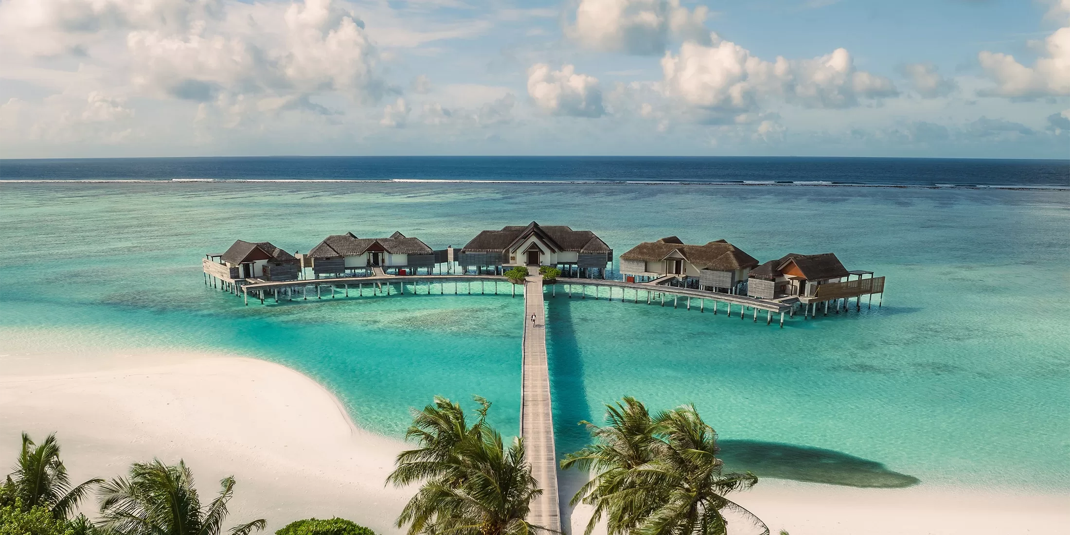 Aerial view of a picturesque beach resort with overwater bungalows forming a semicircle in turquoise waters. A long wooden walkway connects them to the sandy shore, surrounded by palm trees under a partly cloudy sky.