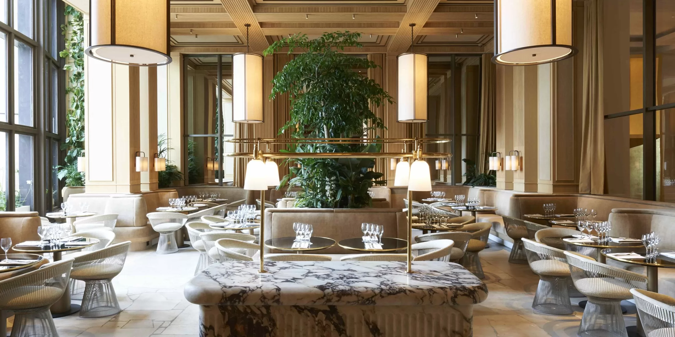 Elegant restaurant interior with a central marble table, surrounding tables set with glassware, modern wooden ceiling, large hanging lamps, and a tall plant against mirrored walls, creating a sophisticated ambiance.