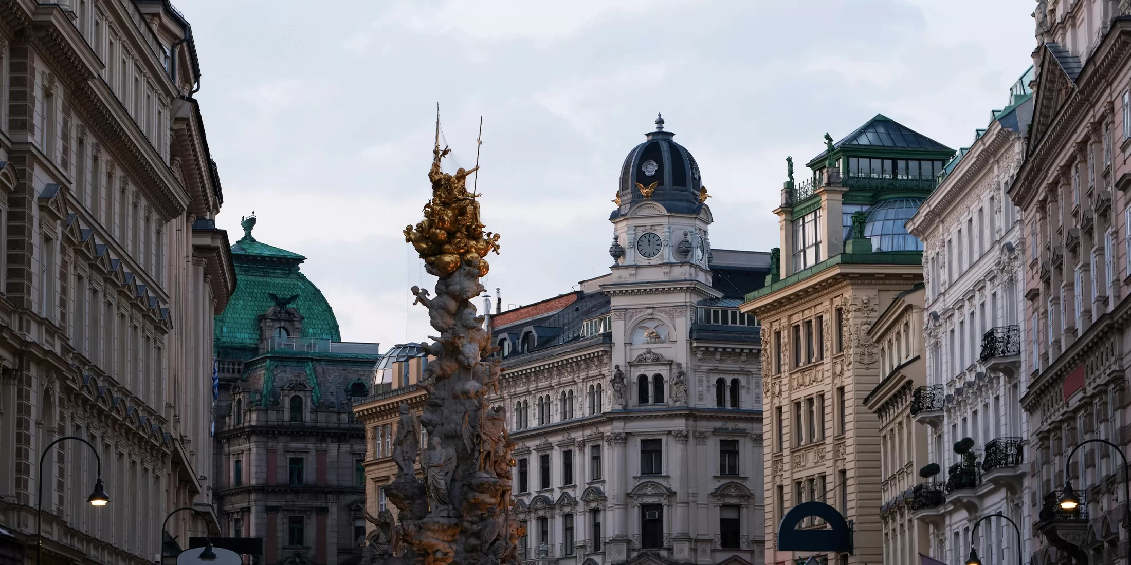 A historic European street scene featuring ornate buildings with intricate facades and green roofs. A prominent column with gold statues stands in the center. The sky is overcast, adding to the old-world charm.