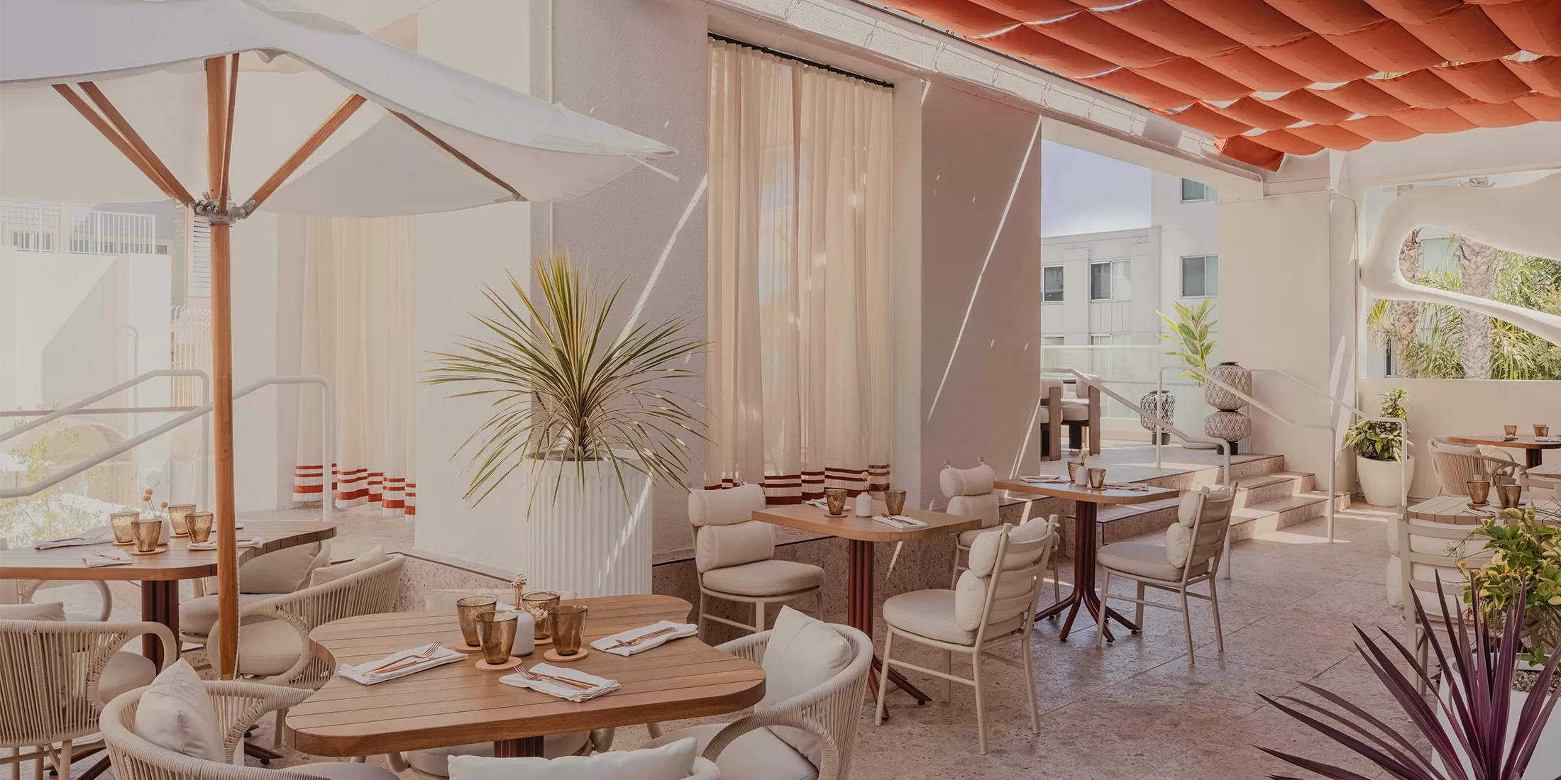 Outdoor restaurant patio with wooden tables, cushioned chairs, and white umbrellas. Potted plants decorate the space, while soft lighting and curtains create a cozy atmosphere. A red awning and building in the background complete the scene.