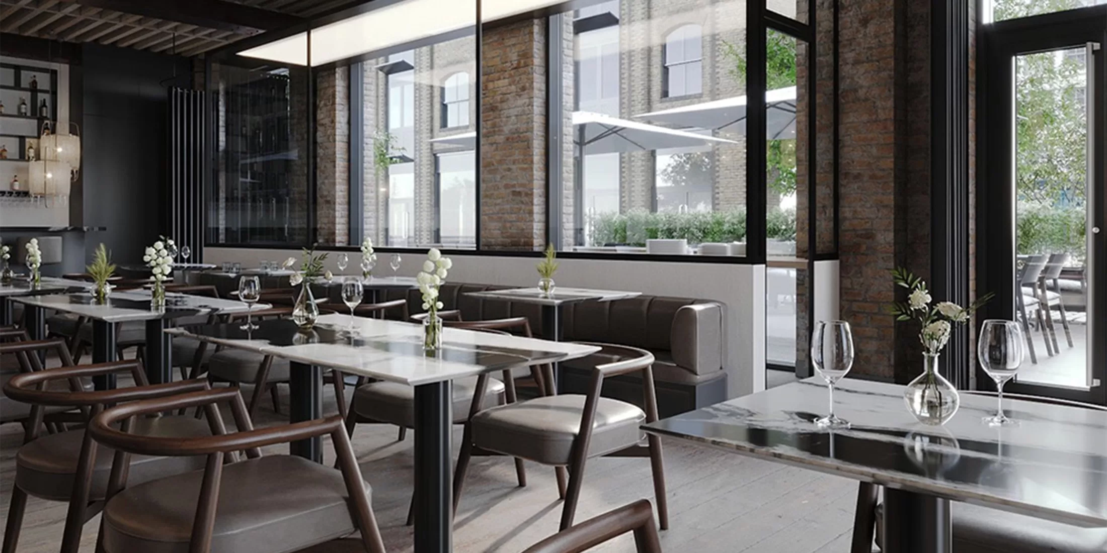 Modern restaurant interior with sleek tables, wooden chairs, and large windows letting in natural light. Small vases with flowers and neatly set wine glasses decorate each table. Brick walls and outdoor greenery visible.