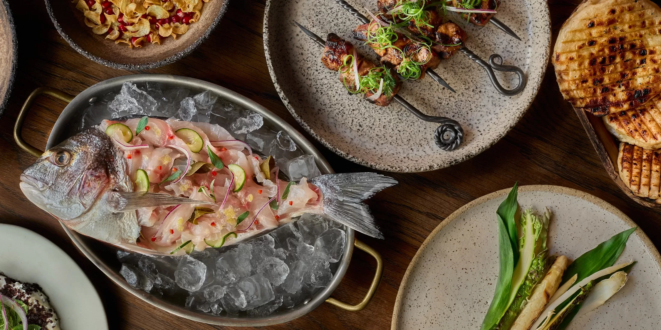 A whole raw fish sliced thinly and served on ice with lime, surrounded by plates of grilled skewers, flatbread, greens, and a salad, all arranged on a wooden table.