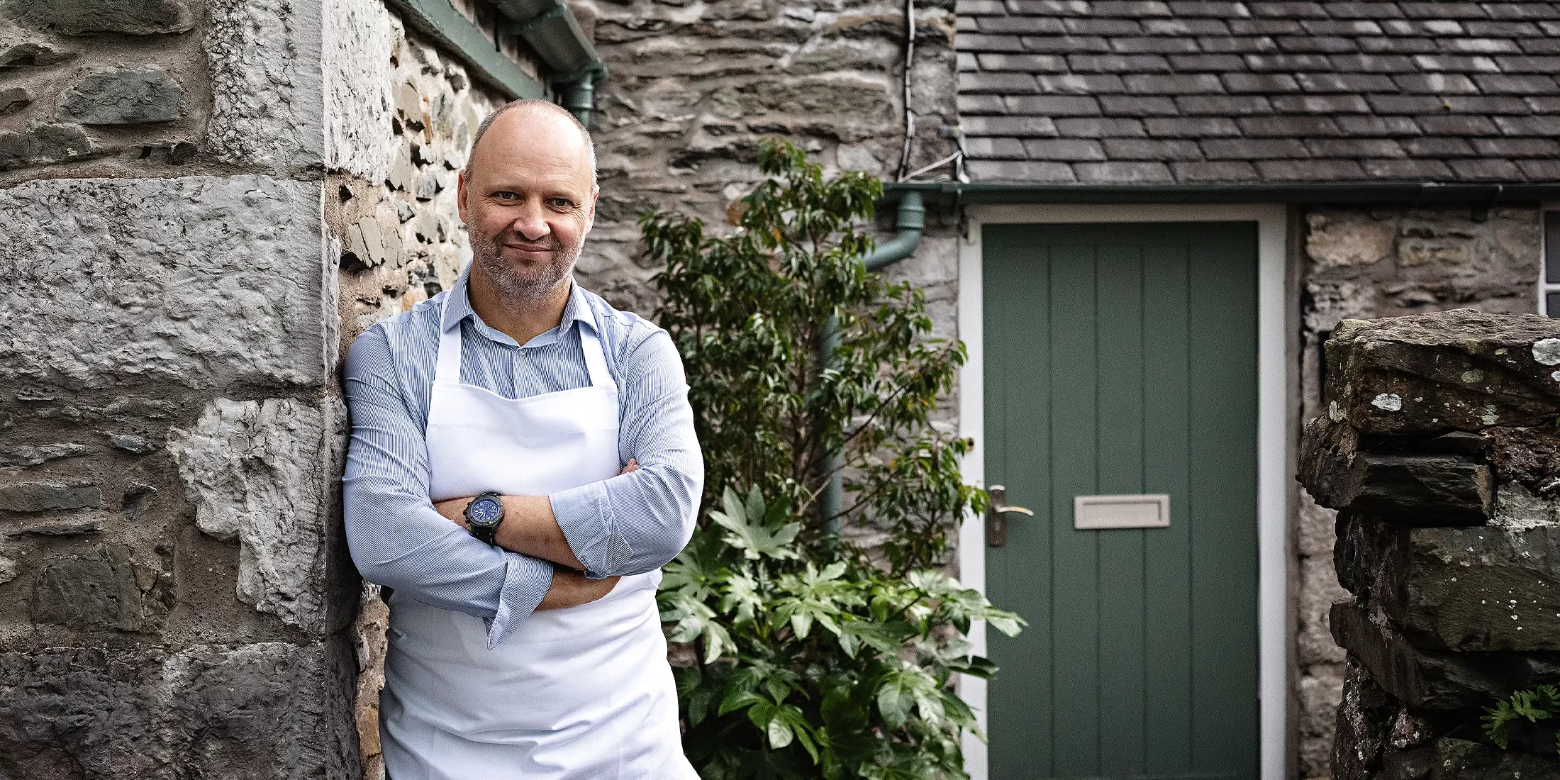 Chef Simon Rogan wearing a white apron and blue shirt, standing with his arms crossed, smiling, against a stone wall. Behind him, there is a green door and plants, with more stone walls in the background.