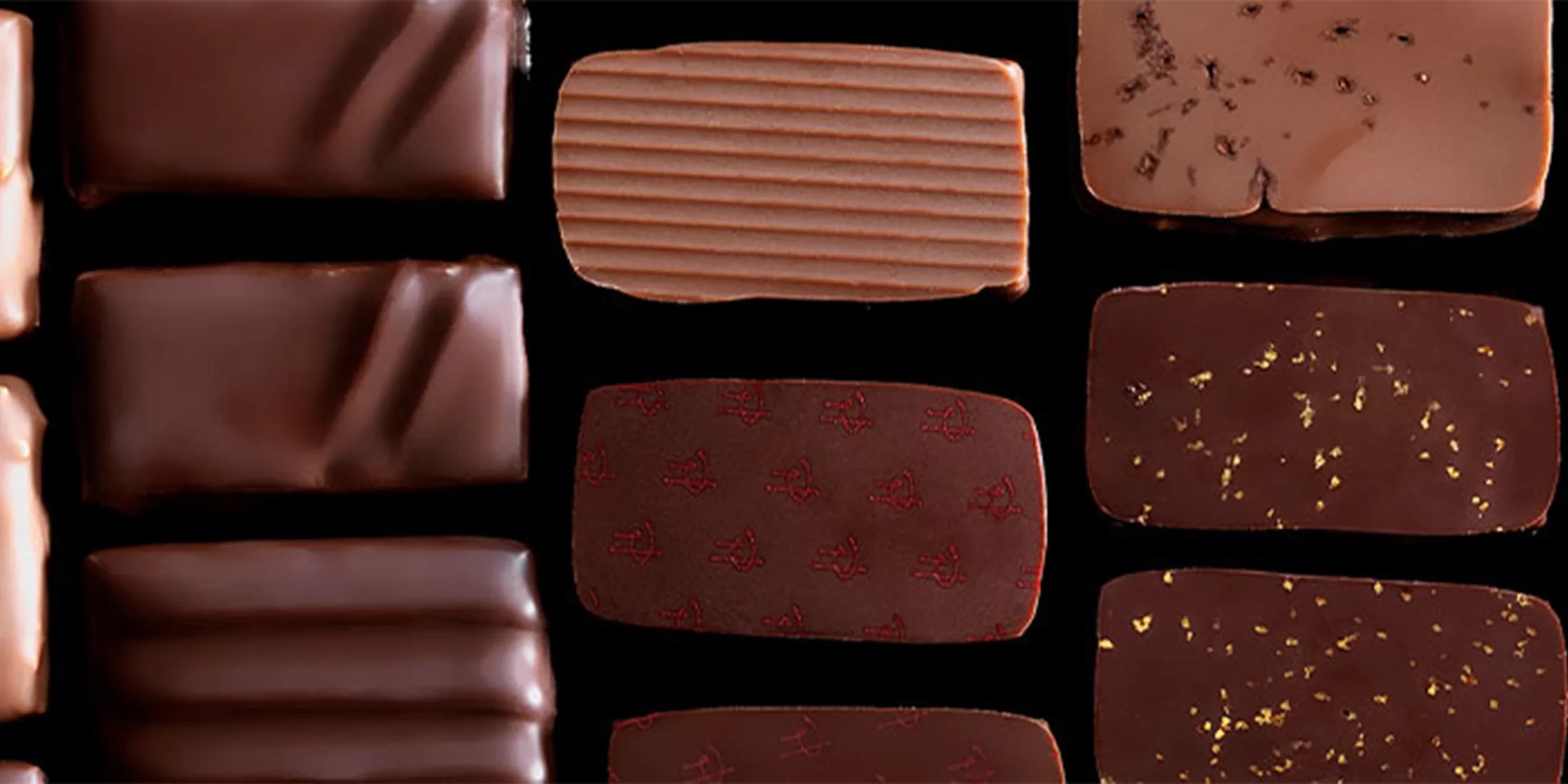 A selection of assorted chocolates arranged in neat rows on a black background, featuring various shapes, textures, and decorations such as gold flakes and patterned surfaces.