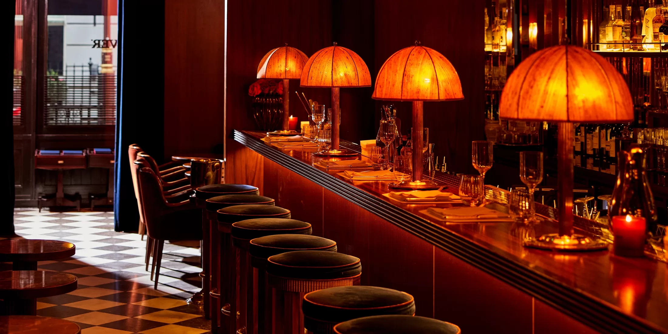 A dimly lit bar with vintage-style orange lamps, velvet-topped stools, and a checkered floor. Table settings are neatly arranged on the bar counter, creating an inviting, cozy atmosphere.