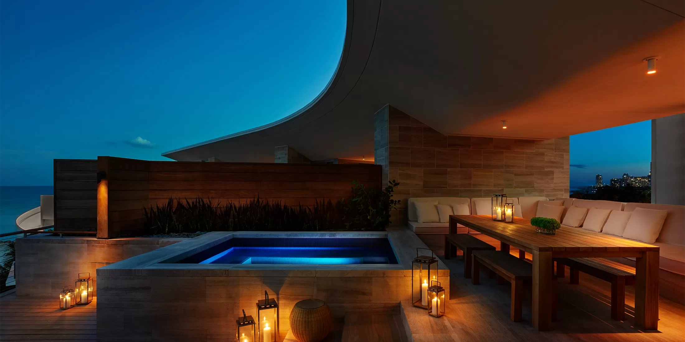 A modern outdoor patio at dusk features a small illuminated pool, wooden dining table with cushioned seating, and lanterns providing warm light, with an ocean view in the background.