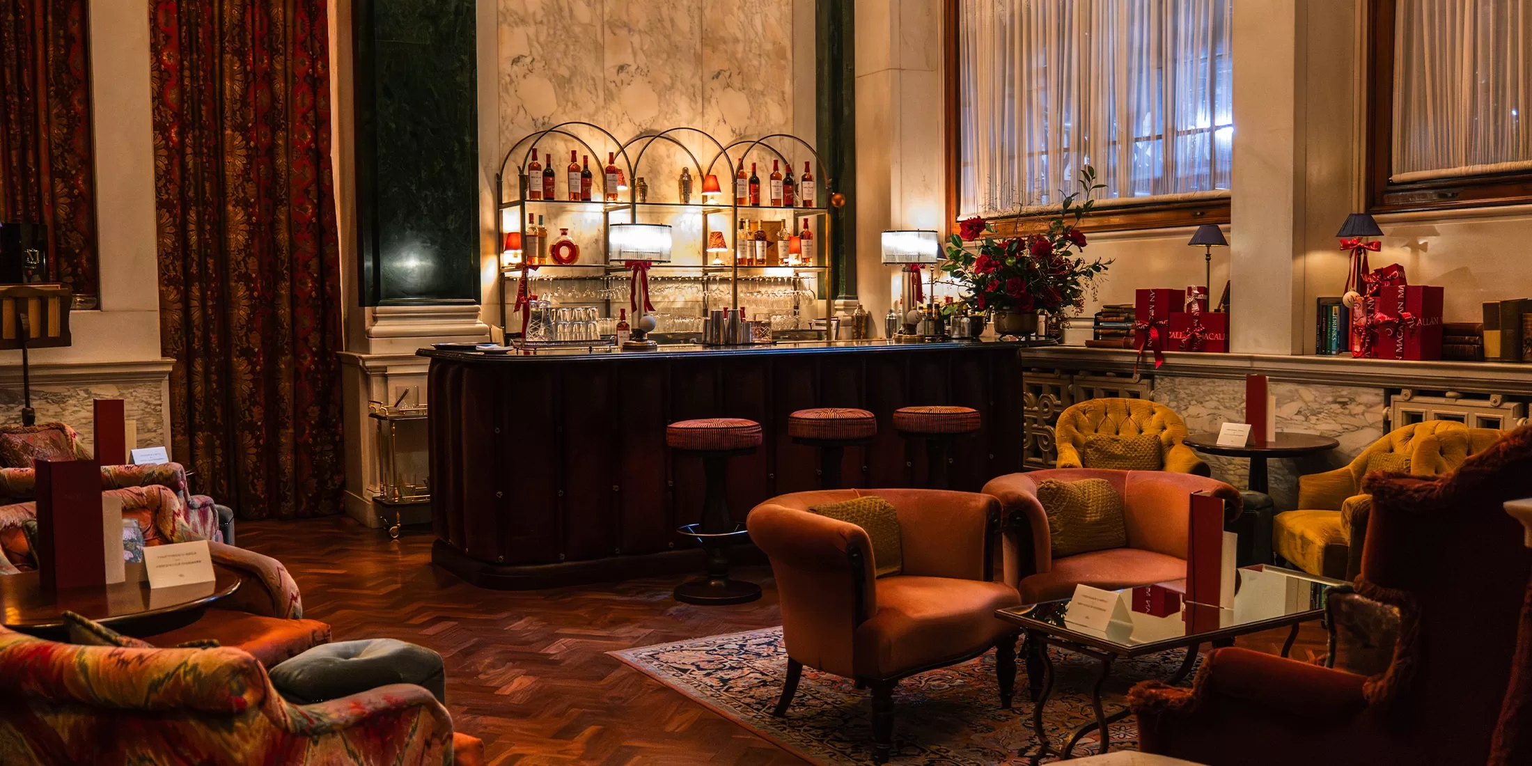 A cozy, elegant lounge with plush armchairs, a patterned rug, and a marble-topped bar lined with stools. Warm lighting, rich drapes, and an array of bottles create a luxurious, inviting atmosphere.