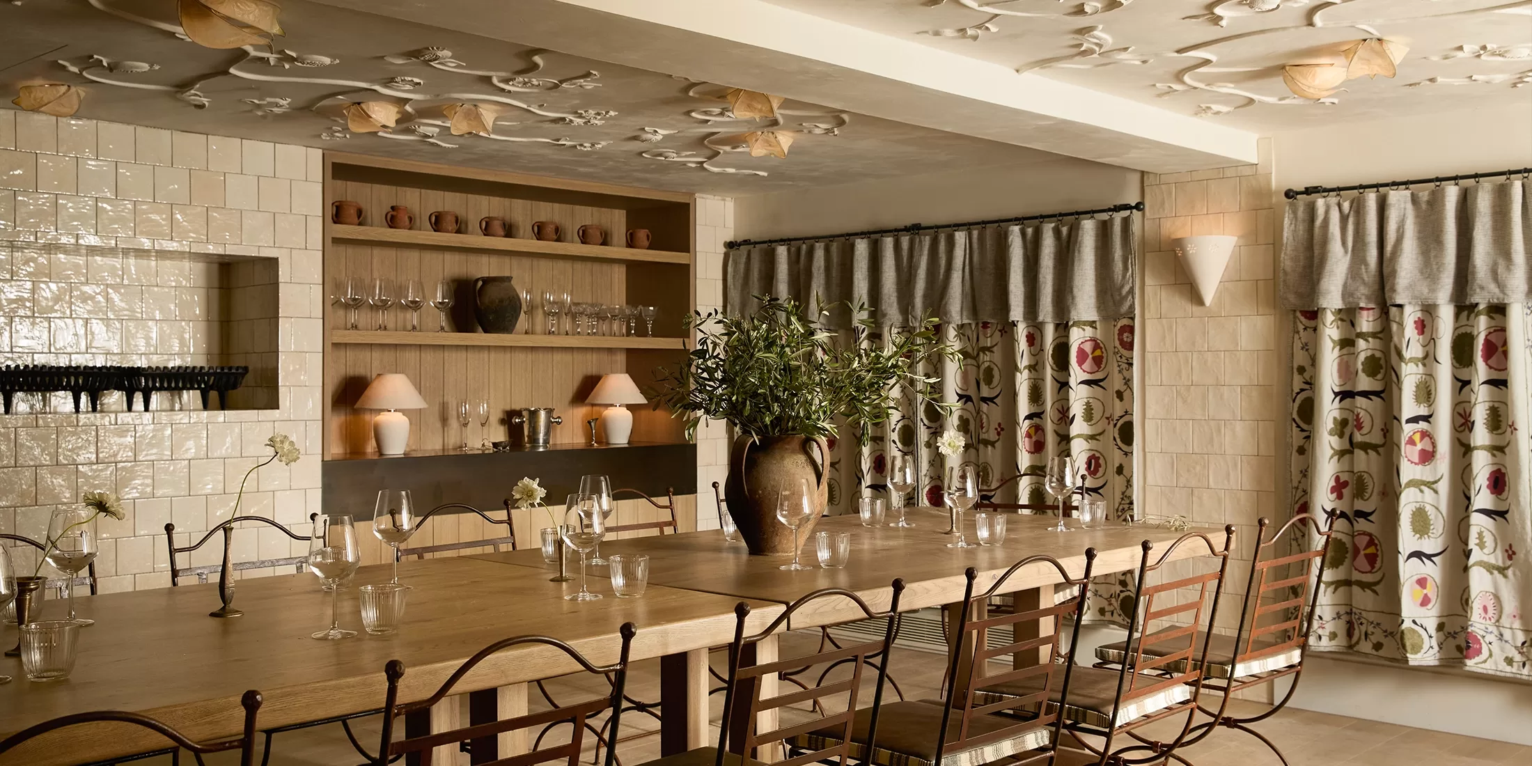 A cozy dining room with a long wooden table, metal chairs, an ornate ceiling, a large vase with greenery, shelves holding crockery, and patterned curtains hanging by the windows. Warm, soft lighting creates an inviting atmosphere.