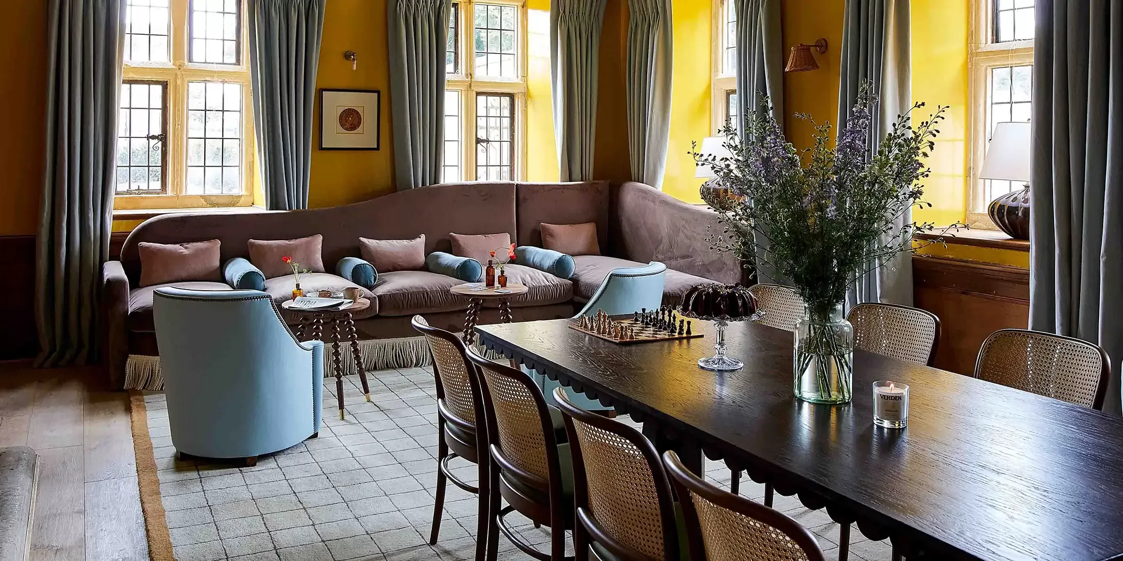 A cozy living room with yellow walls, large windows, a brown sectional sofa with blue and pink cushions, blue armchairs, a dark wooden dining table with cane chairs, and a vase of flowers on the table.