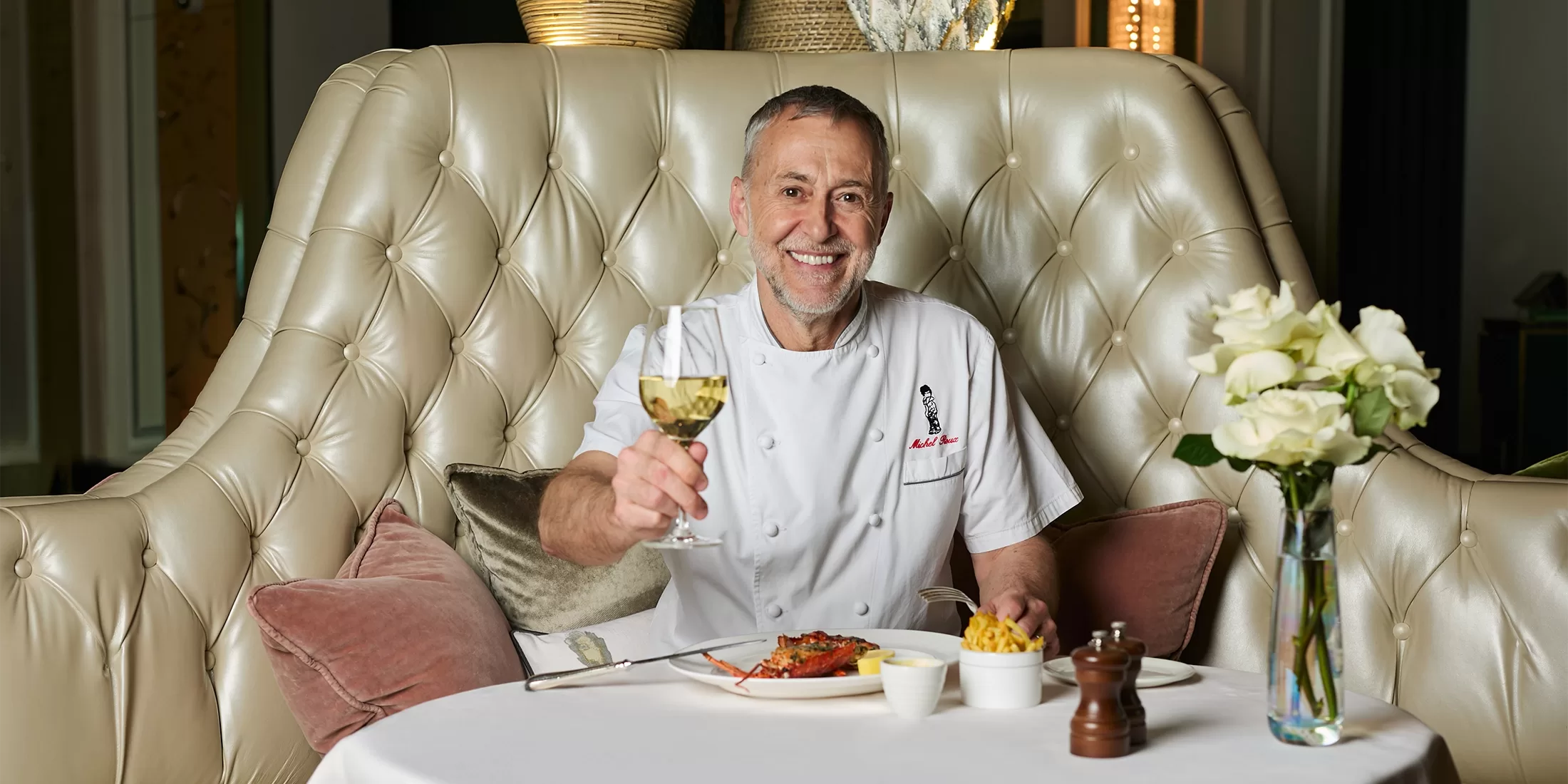 Michel Roux sat at a tall leather back quilted dining booth, smiling with a glass of white wine in hand and a table full of food at Chez Roux at The Langham, London