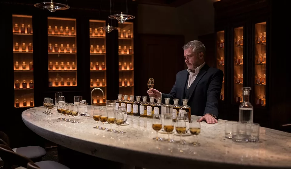 A man in a suit stands at a marble bar, examining a glass of whiskey. Small bottles and glasses filled with whiskey are arranged on the counter, with shelves of backlit bottles lining the dim room.
