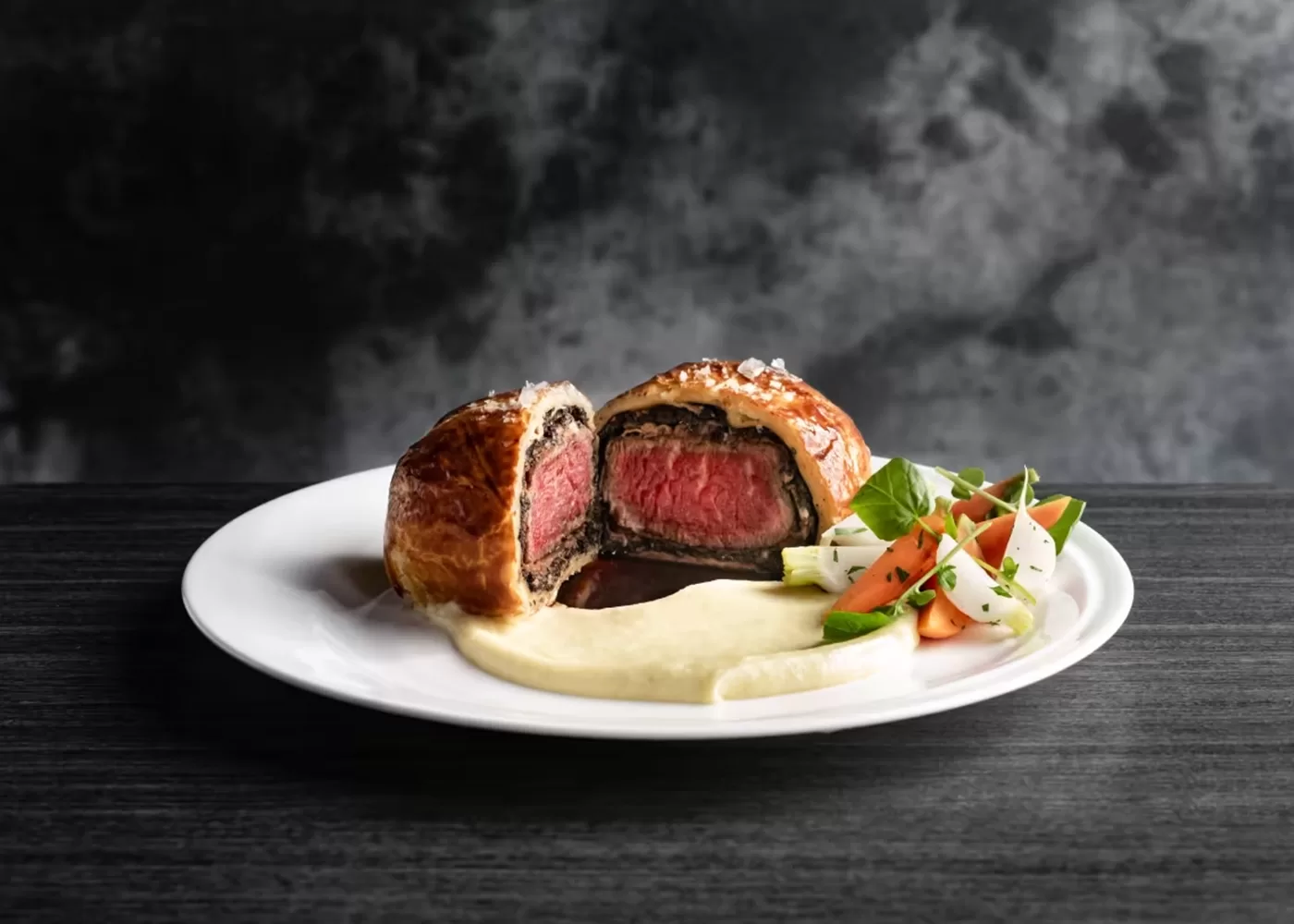 A plated Beef Wellington sliced open to reveal a medium-rare center, served with creamy mashed potatoes and a garnish of mixed vegetables and herbs on a white plate against a dark background.