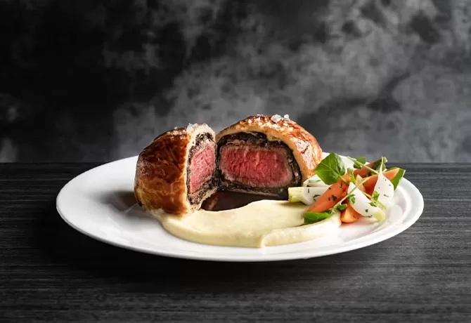A plate with sliced beef Wellington showing a pink center, served with creamy mashed potatoes and a side of mixed vegetables, against a dark, textured background.