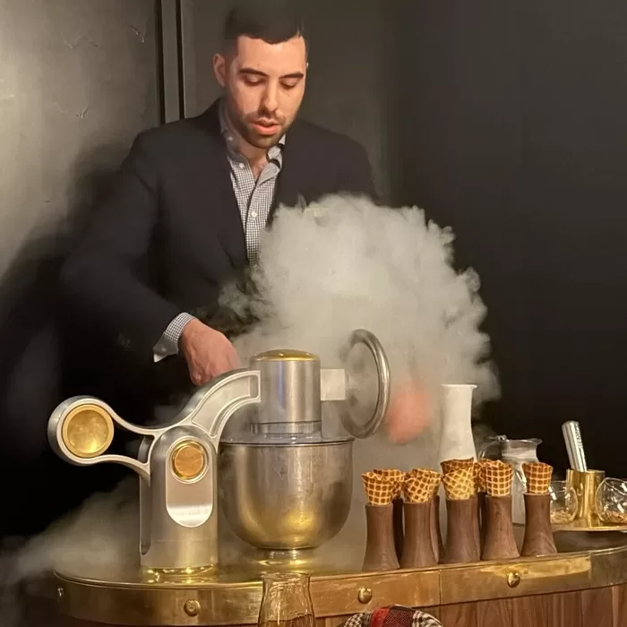 A man in a suit prepares ice cream using a stand mixer and liquid nitrogen, creating a cloud of vapor. Waffle cones are arranged on a marble-topped counter beside him.
