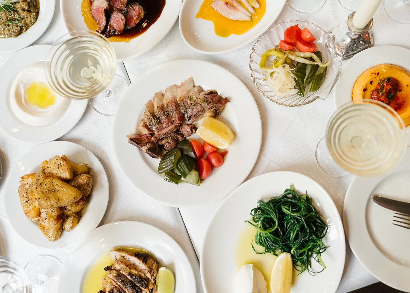 A table set with various plated dishes, including sliced meat with lemon and vegetables, sautéed greens, roasted potatoes, pickled vegetables, and glasses of white wine on a white tablecloth.