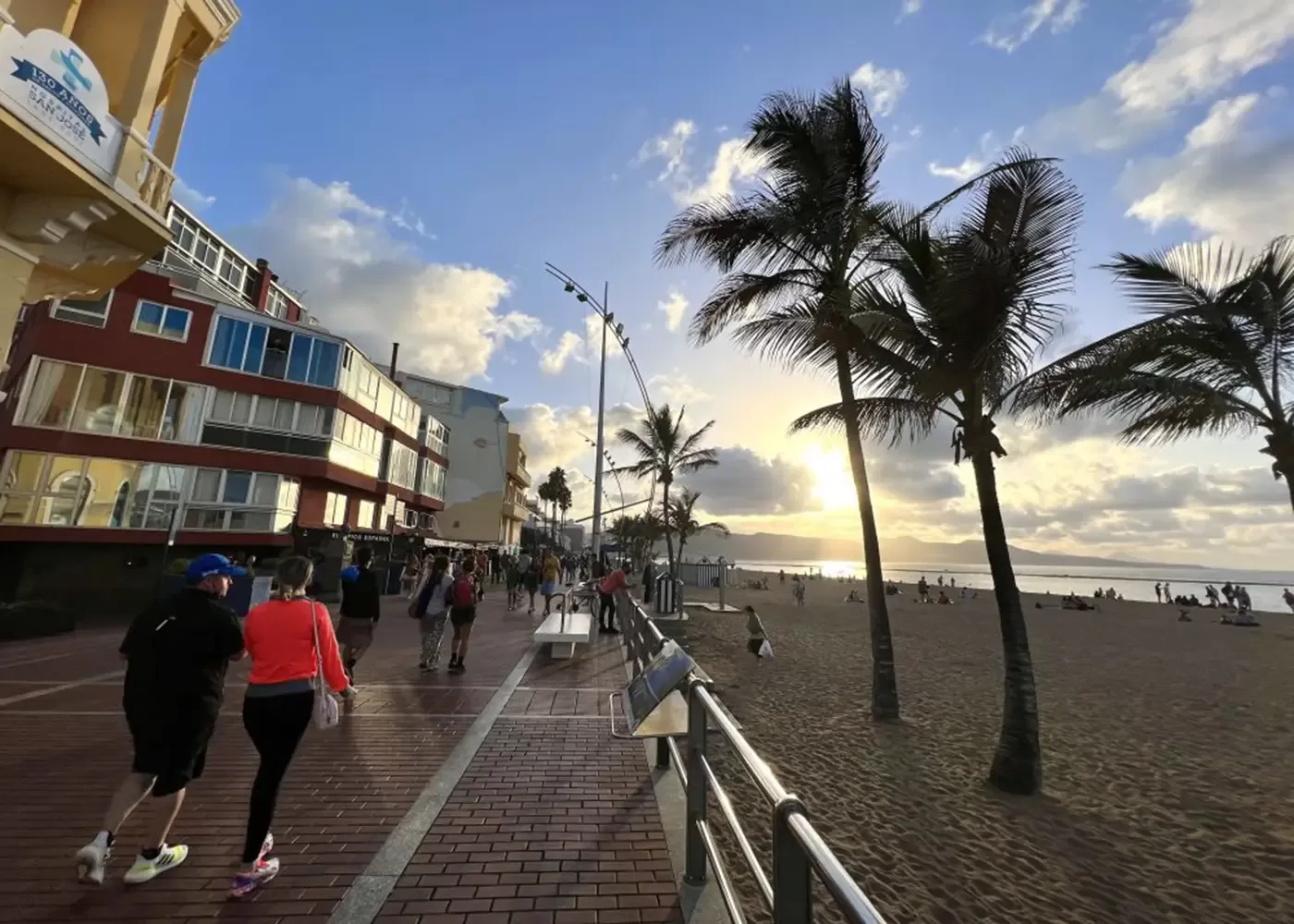 People walk along a beachside promenade at sunset, with palm trees, sandy beach, and buildings on the left. The sky is partly cloudy, and the sun is low on the horizon, casting a warm glow over the scene.