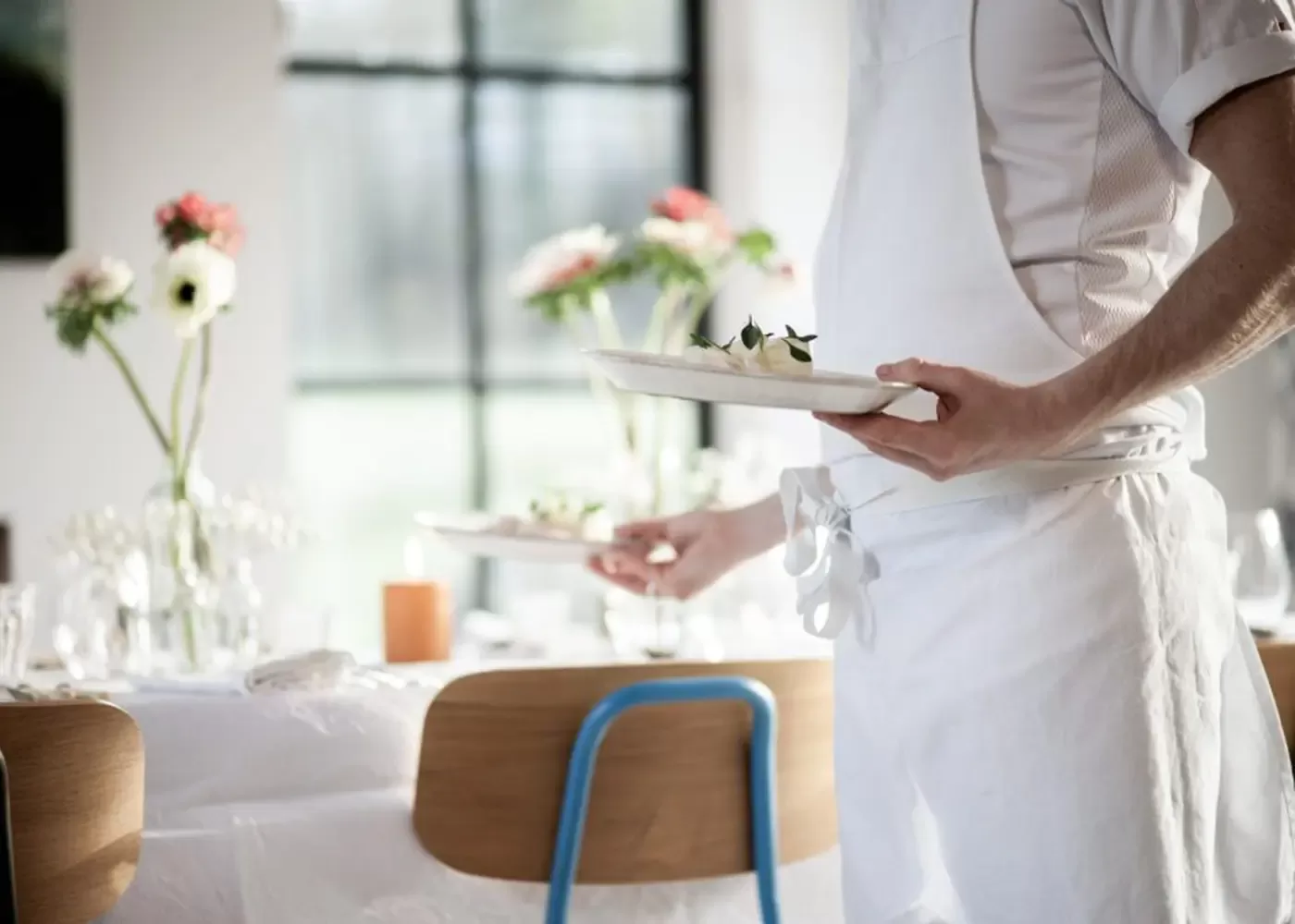 A person wearing a white apron serves plates of food in a bright, elegant dining setting with a white tablecloth, wooden chairs, and vases of flowers on the table.