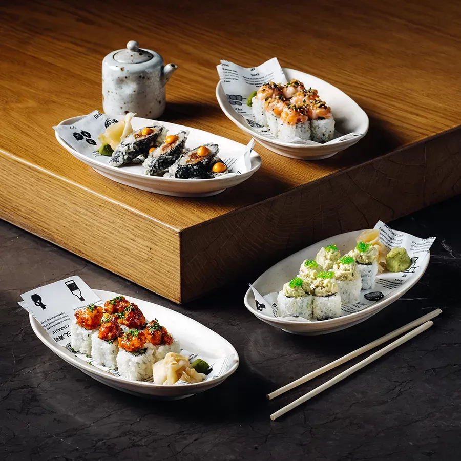 Three plates of assorted sushi rolls with various toppings are arranged on a dark table with chopsticks, next to a white teapot on a wooden surface in the background. Each plate includes slices of pickled ginger.