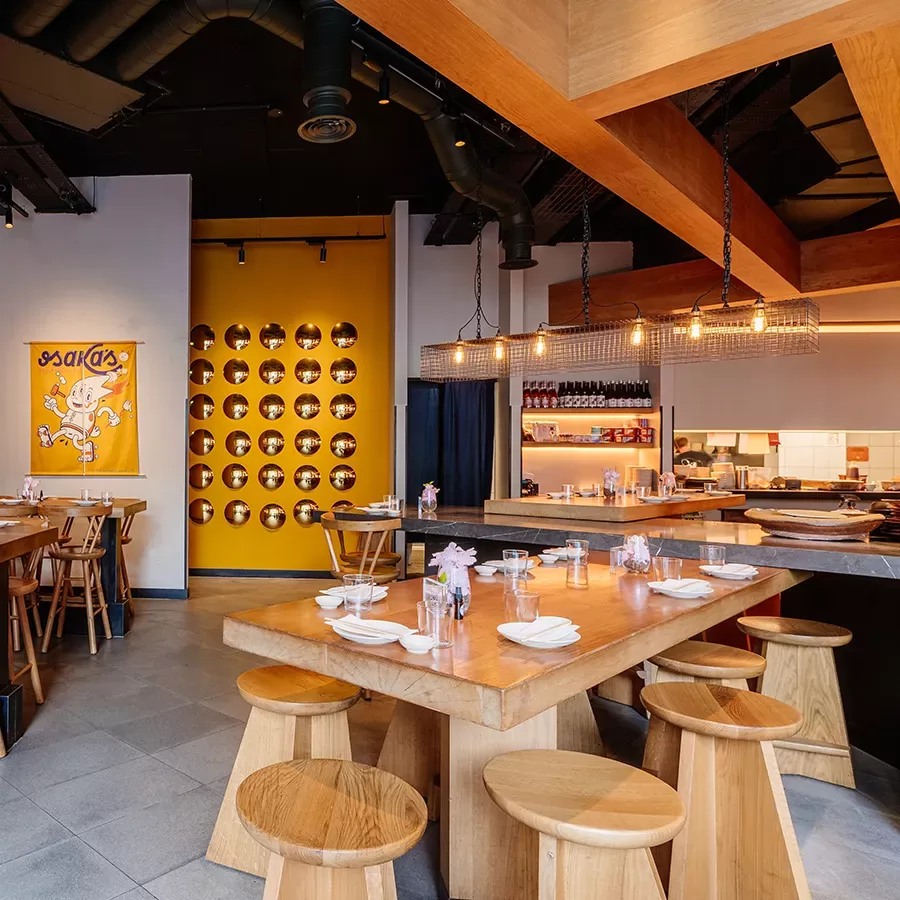 Modern restaurant interior with wooden tables and stools, neatly set with plates and glasses. A yellow wall with circular cutouts and a colorful artwork add character, while warm pendant lights create a cozy ambiance.
