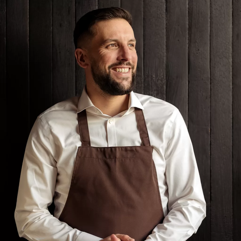 A man with short dark hair and a beard, wearing a white shirt and brown apron, stands smiling and looking to the side in front of a dark wooden wall.