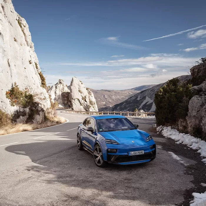 A blue sports car is parked on a winding mountain road surrounded by rocky cliffs and sparse vegetation, with patches of snow and a clear sky in the background.
