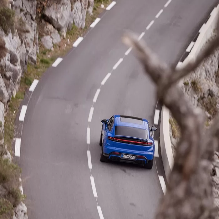 A blue sports car drives along a winding mountain road bordered by rocky cliffs and vegetation, viewed from above.