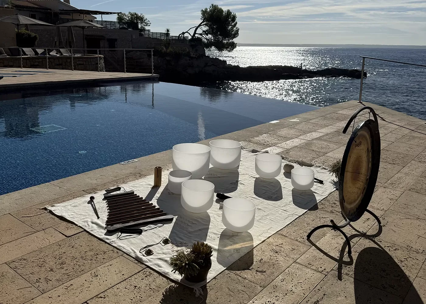 A white cloth with crystal singing bowls, a gong, mallets, and other musical instruments is set up by an infinity pool overlooking the ocean on a sunny day.