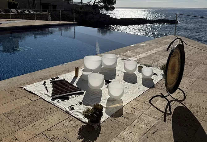 A set of white crystal singing bowls, a gong, chimes, and other sound healing instruments are arranged on a white cloth beside a pool, overlooking a calm sea under sunlight.
